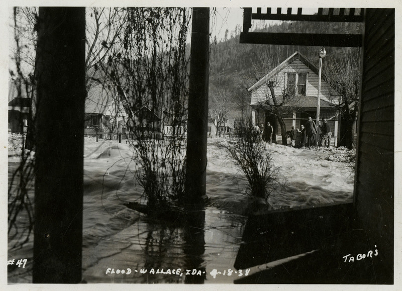 Water flows over the surface of a porch during a flood in Wallace, Idaho. There are also a few trees in frame and a small group of people on nearby dry land.