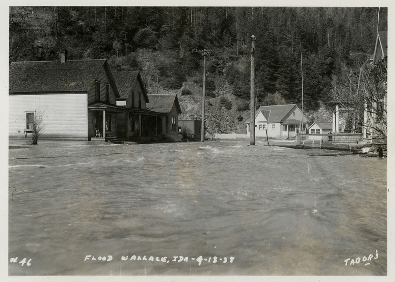 Water rushes past several buildings during a flood in Wallace, Idaho. There are a few telephone poles in view.