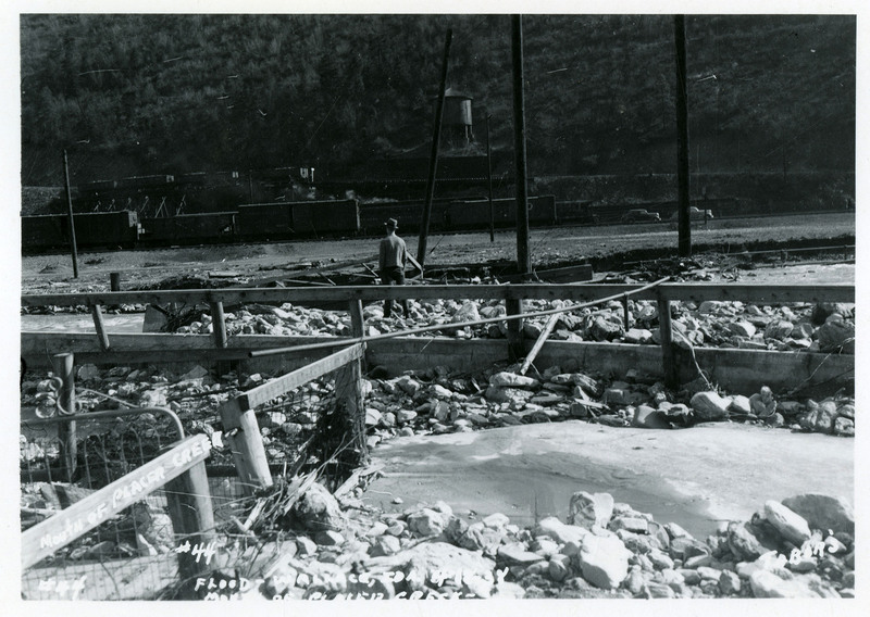A man stands on top of flood debris while facing away from the camera. He appears to be looking at the river. There are several damaged fences in view and a few cars in the background.