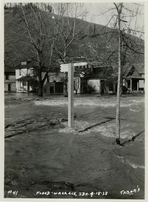 Water surrounds the base of a signpost during the Wallace flood. There are a few trees and buildings in view. The signpost points to Spokane, Coeur d'Alene, Kellogg, Avery, Yellowstone Park, and Missoula, and lists the distances to those locations from the location of the signpost.
