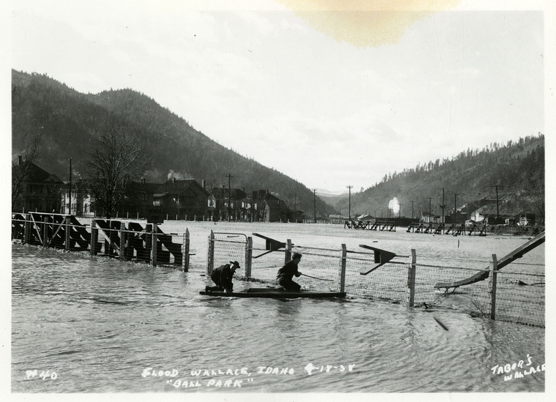 Two people kneel on wooden boards near the fence of Ball Park in Wallace, Idaho during the flood. The ground is almost entirely covered in water.