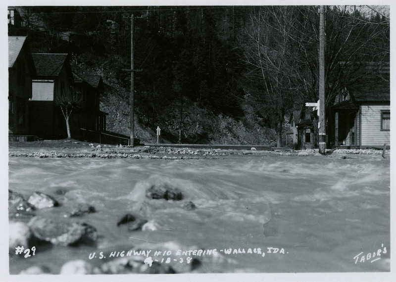 Water rushes past several buildings during a flood in Wallace, Idaho. Photograph is labeled as "U.S. Highway #10 Entering." There are a few telephone poles in view.