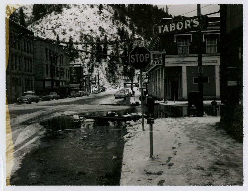 Water covers a street in a commercial area of Wallace during the Christmas Flood. Tabor's, Sweets Hotel, and several bars are in view and several cars are parked on the side of the road.