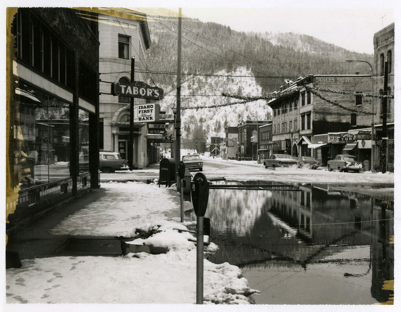 Water covers a street in a commercial area of Wallace during the Christmas Flood. Tabor's, Idaho First National Bank, and other nearby businesses are in view and several cars are parked on the side of the road.