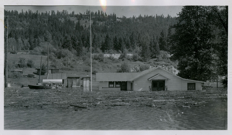 Placer Creek overtakes a building during the Wallace flood. A boat can be seen in the background and debris is strewn across the surface of the water.