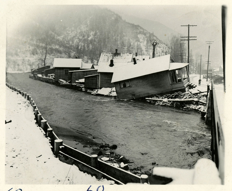 A river encroaches on several buildings during the Wallace flood. A least one of them appears to have sustained structural damage and appears to be falling into the river. There is snow on the ground and a bridge nearby. There is a person and several telephone poles in the background.