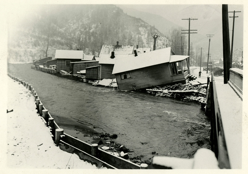 A river encroaches on several buildings during the Wallace flood. A least one of them appears to have sustained structural damage and appears to be falling into the river. There is snow on the ground and a bridge nearby. There is a person and several telephone poles in the background.