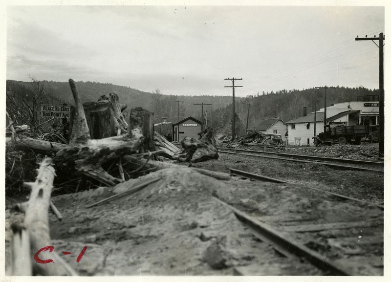 Downed trees and other plant debris litter a train track after the Wallace flood. Several buildings and telephone poles are visible in the background, as well as a sign that says "Place No Cars This Point [unintelligible]." 