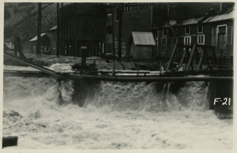 Water flows near buildings during the Wallace flood. Water appears to be falling from a slightly higher elevation to a lower elevation on the street.