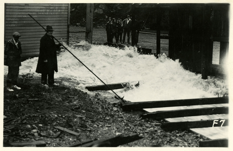 Two groups of people stand on opposite sides of rushing floodwaters during the Wallace flood. One man in the foreground appears to be holding a long pole.