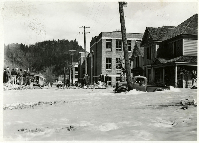 Several people observe floodwaters from spots of dry land during the Wallace flood. At least one person is attempting to drive a truck through the waters. There are several buildings, telephone poles, and other vehicles in the background.