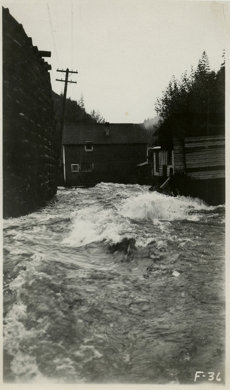 Flood waters rush past several buildings and what appears to be a brick wall during the Wallace flood. A leaning telephone pole is also visible.