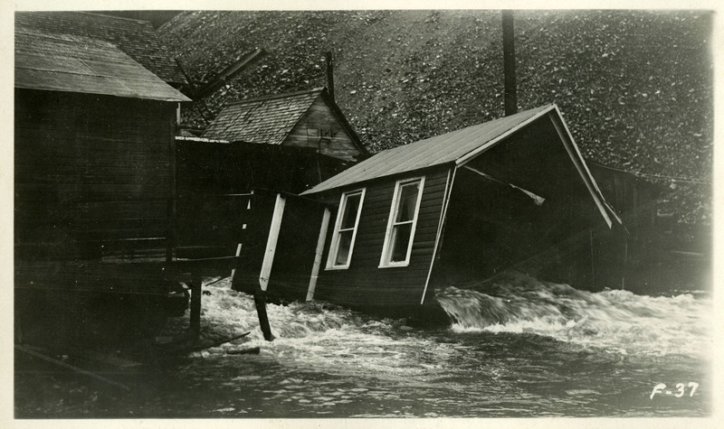 Water rushes through and out the side of a damaged shed-like building during the Wallace flood. Water appears to have reached other nearby buildings as well.