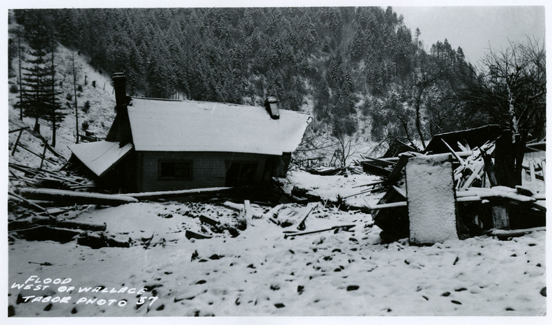 A shot of a heavily damaged house west of Wallace after the flood. Debris is visible in the surrounding area and snow covers most visible surfaces.