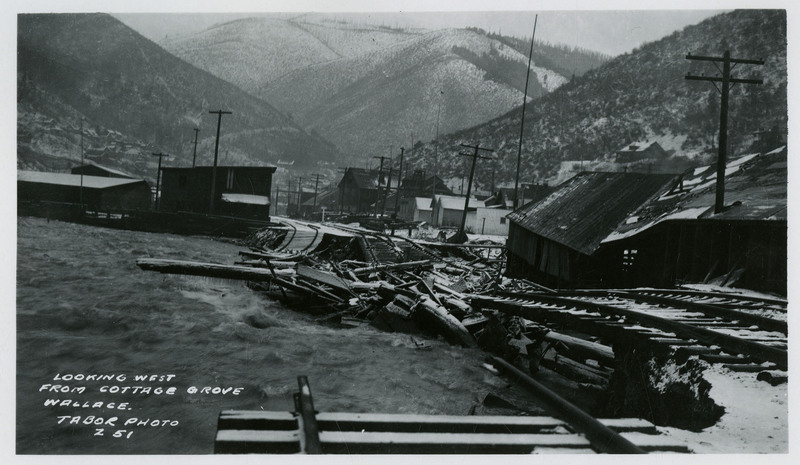 Floodwaters rush underneath damaged railway tracks and buildings during the Wallace flood. The photo labels this viewpoint as looking west from Cottage Grove.