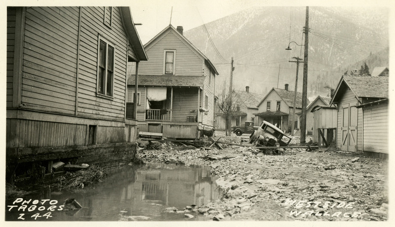 The driver's side door of a car is open and the driver absent after attempting to drive over debris left by the wallace flood. A dog stands near one of the nearby damaged houses and another car drives by on the street in the background. A large puddle takes up most of the foreground.
