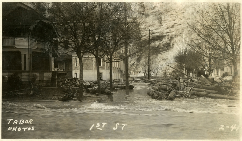 Debris and floodwaters cover First Street during the Wallace flood. A few houses, trees, and telephone poles are visible in the background.