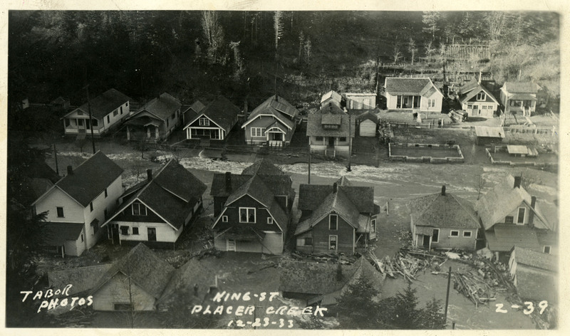 Floodwaters flow between King Street and Placer Creek during the Wallace flood. Several homes appear to be damaged and debris is visible in the foreground.