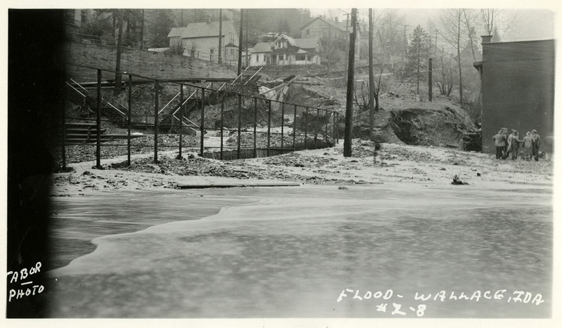 Water pours down a hillside, past damaged infrastructure, to a larger amount of flooding at the bottom of the hill. A group of people appear to be talking to each other on the side of the frame.