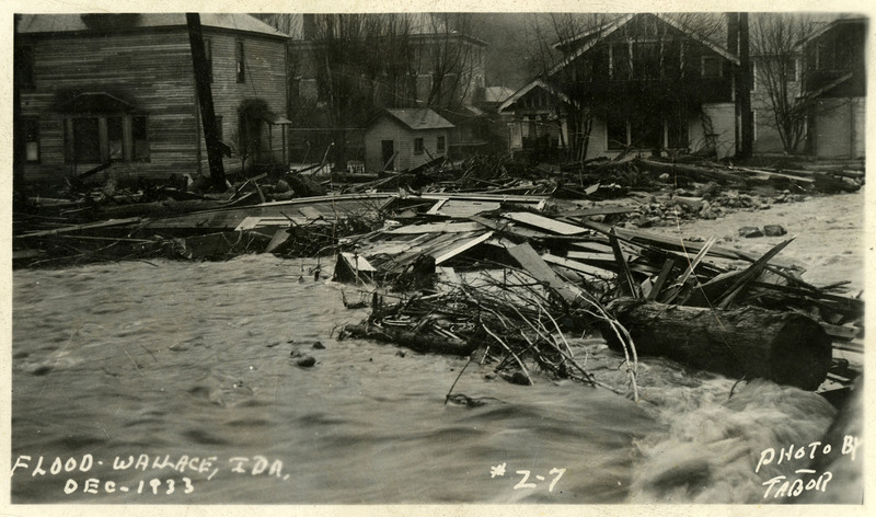 A pile of debris, including wood and metal, impedes rushing floodwaters during the Wallace flood. There are several buildings in the background.