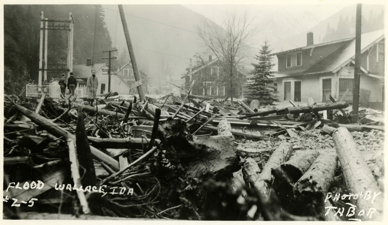 Three people stand on a pile of debris following the Wallace flood. A telephone pole standing in the middle of the pile appears to be damaged. Several buildings are visible in the background.