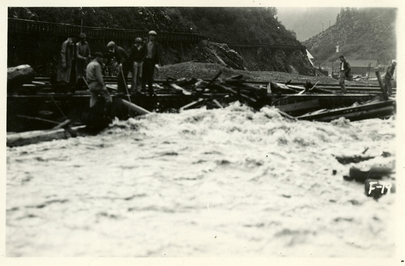 A group of seven people stand on a pile of structural debris during the Burke Canyon flood. A few buildings are visible in the background.
