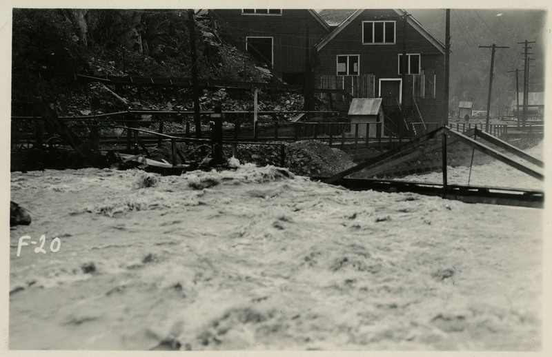 A river of rushing water almost meets street level during the Burke Canyon flood. Several buildings are visible in the background.