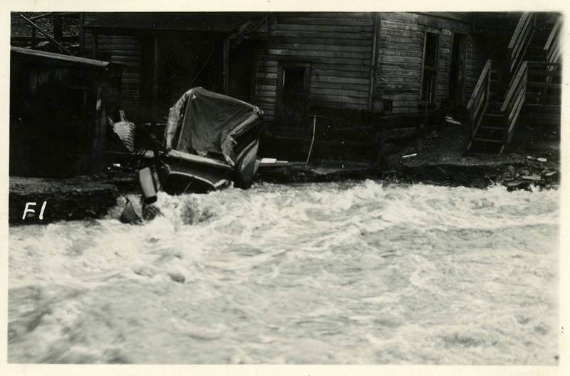A damaged car is visible on a small debris pile near a building during the Burke Canyon flood. Water continues to rush through the surrounding area.