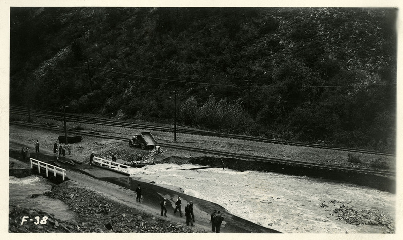 Flooding flows between a road and set of railroad tracks during the Burke Canyon flood. A group of people walks along the road in the foreground.
