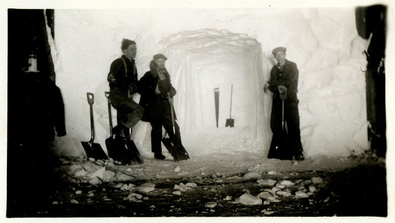 Three people with snow shovels work to remove snow from a mineshaft. A few extra shovels and a saw are leaned up against the snow.