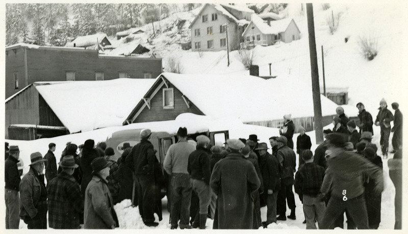 People gather near a vehicle with open rear doors after a snowslide in Burke. Snow covers buildings in the background.