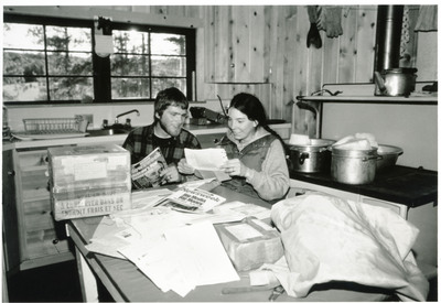 A photo by Pat and Greg Hayward--set up on tripod--No other human within maybe 20 miles. Pat and Greg Hayward at Cold Meadows guard station in the River of No Return Wilderness examining the mail which has accumulated over the past month. During winter mail arrived by ski plane every 2 weeks to a month. (On this day we moved from Chamberlain to Cold Meadows by ski plane. You can see th esnow melting on the stove to begin the supply of water for the 2 week stay at Cold Meadows).