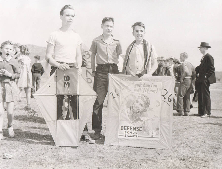 Photo text: 'Winner in a kite flying contest, Boise, Idaho, April, 1942. Sponsored by the Recreation department of Idaho WPA, and the Idaho Daily Statesman.' Three boys stand with two kites. The kite on the right advertises defense bonds. Note: This image is part of a Work Progress Administration publicity series.