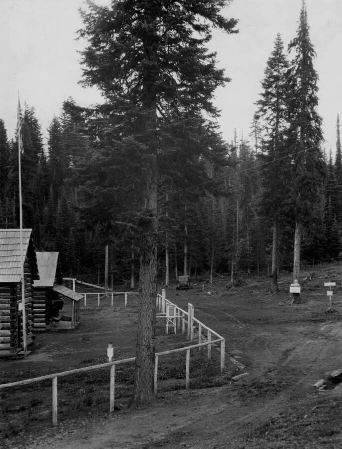 The driveway area of Adams Ranger Station. Pole fencing runs alongside the drive way along with what is likely a weather data collection box near the center of the photo. Two log building including the main building sit to the left of the photo. A car can be seen in the background.