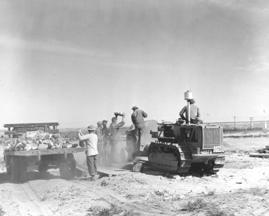 A group of CCC workers from Mountain Home Camp G-99 work to remove excess soil. A large ditch is dig that a truck can pass through. A platform is build over with the grinder on top of it. Grind from large rocks falls through hole in platform directly in bed of truck below. Image part of CCC-Idaho Indian Division.