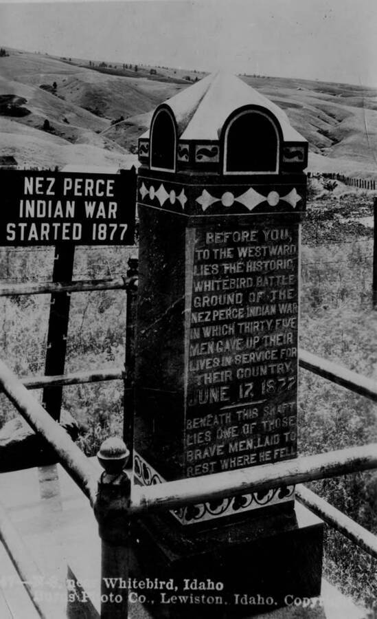 Photo caption on back: 'Photo of graves of soldiers and civilians killed in the Nez Perce Indian War. Secured by R.H. Fletcher from Aaron F. Parker, Grangeville, Idaho.' Sign to left reads: Nez Perce Indian War started 1877. Monument reads: Before you to the Westward lies the historic Whitebird battle ground of the Nez Perce Indian War in which thirty-five men gave up their lives in service for their country June 17, 1877. Beneath this shaft lies one of those brave men, laid to rest where he fell.