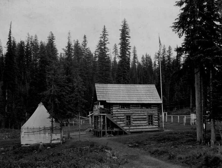 The two-story log main building of Adams ranger station sits among pole fencing. A second plank and tent building sits nearby. The edge of a third log building can be seen behind the main building.