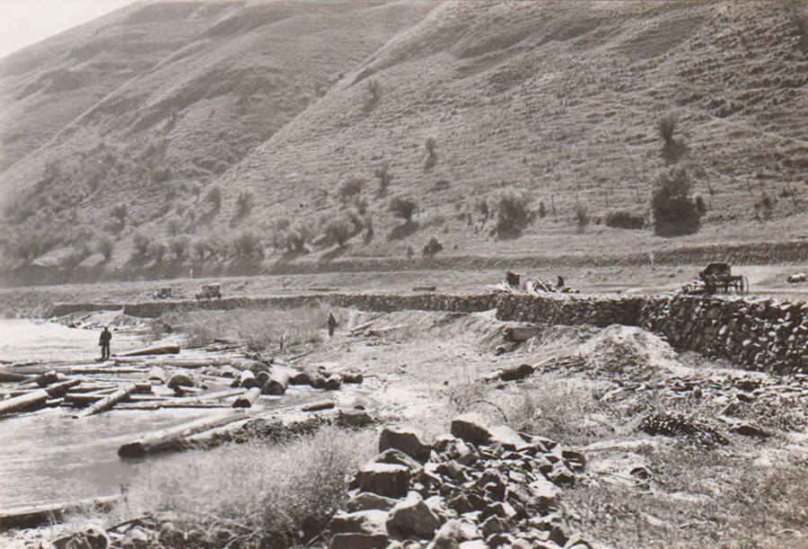 Rip-rap is installed to prevent erosion of banks on the Clearwater River. A man stands on logs in the midground. These log are likely from river log drives where logs were floated down the river to the mills. Logs often collected near the Nez Perce Boom Grounds at this bend. Note: This image is part of a narrative pictoral report to accompany quarterly enrollee program report.