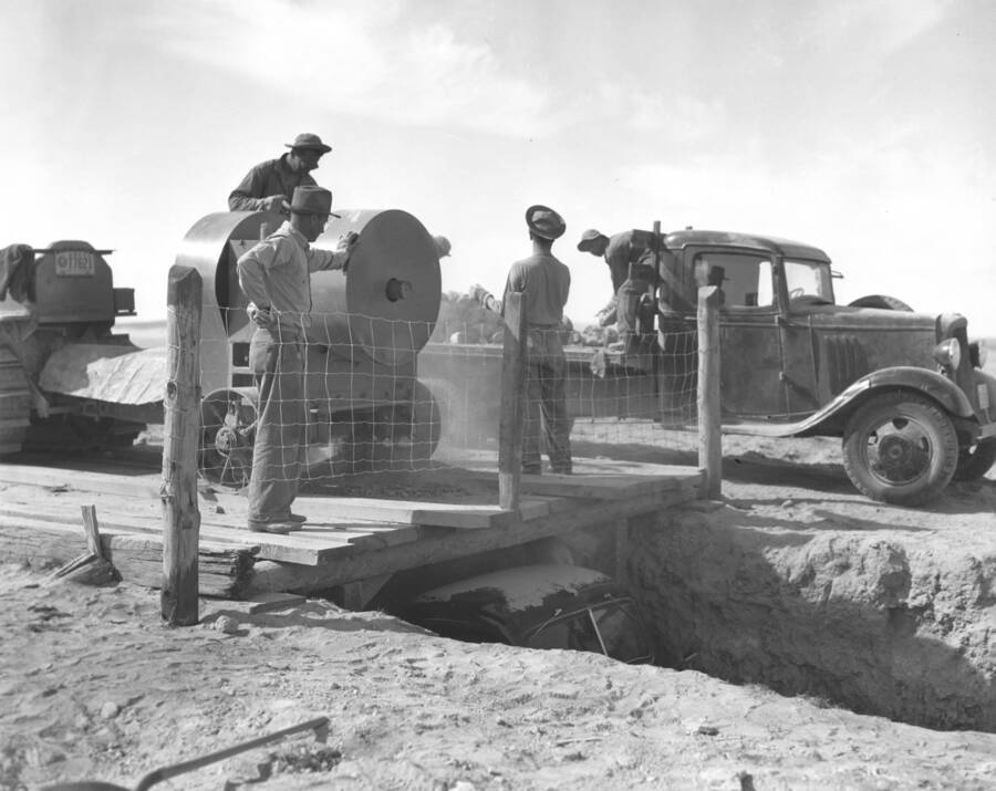 A group of CCC workers from Mountain Home Camp G-99 work to remove excess soil. A large ditch is dug that a truck can pass through. A platform is build over with the grinder on top of it. Grind from large rocks falls through hole in platform directly in bed of truck below. Image part of CCC-Idaho Indian Division.