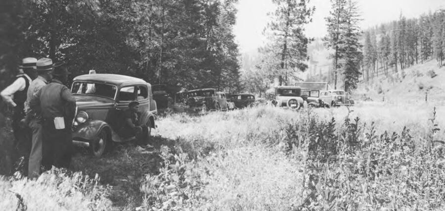 Photo caption: 'This crowd arrived for the picnic and inspection.' A line of cars and are park along forest edge. This image is part of a report regarding farm organizations among tribes in Northern Idaho and the CCC-Indian Division.