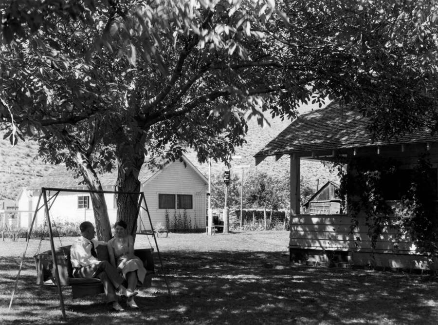 A man and a woman sit in a swing bench outside the Riggins Ranger Station (right) under s tree. A second period structure and garden are in the left side background. Older structures sit farther back and to the right.