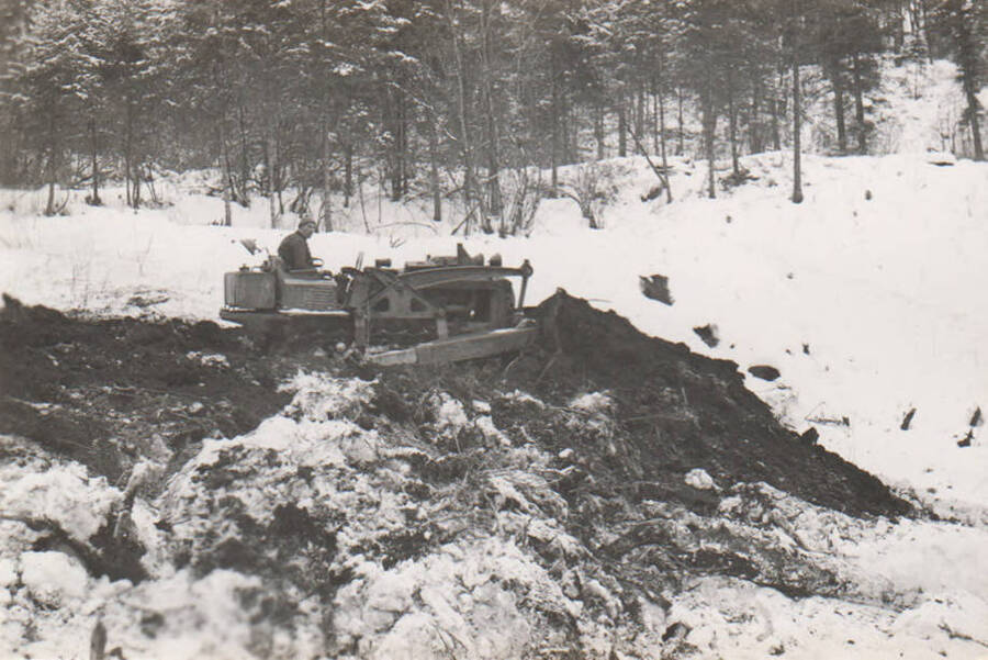 A man bulldozes for trail construction in winter with some inches of snow on the ground, near Fords Creek now known as Jim Ford Creek. Note: This image is part of a narrative pictoral report to accompany quarterly enrollee program report.