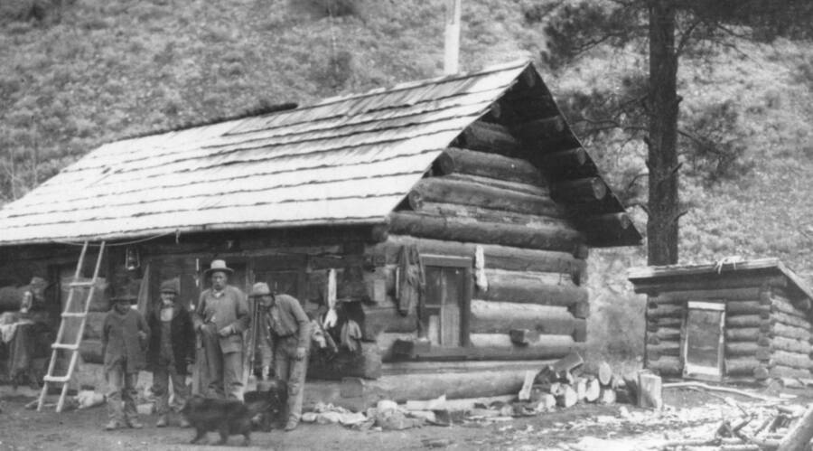 Photo text: 'Sater Cabin circa 1918 - 1928. Man on right is Mike Hogan, others unidentified. Men were reshingling the roof with split shakes. Note blanket used for door of west (left) room and the smokehouse at the right of the cabin. Photo courtesy of Daisy Tappan.'