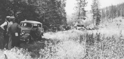 Photo caption: 'This crowd arrived for the picnic and inspection.' A line of cars and are park along forest edge. This image is part of a report regarding farm organizations among tribes in Northern Idaho and the CCC-Indian Division.