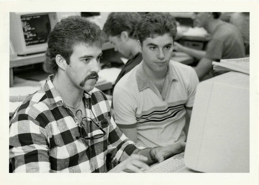 Students sit at computers in the computer lab. Two men sit at one computer.