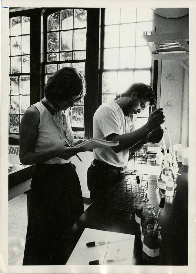 A man and a woman working with scientific equipment. The man is operating the equipment while the woman reads directions or takes notes.