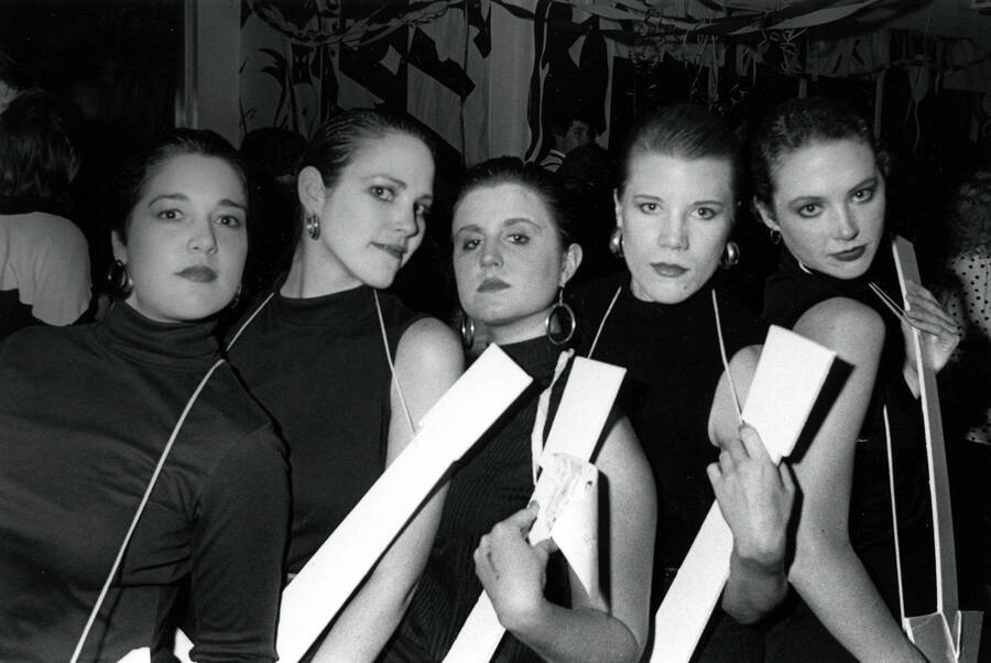 Five women in costume and holding cardboard guitars, very possibly imitating women from Robert  Palmer's ""Addicted to Love"" music video.