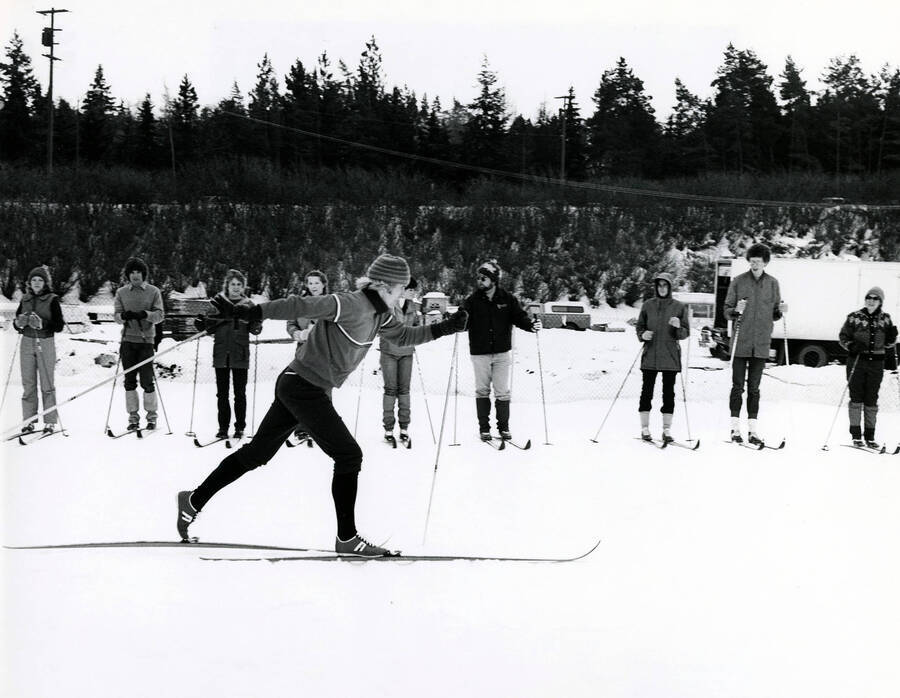The caption on the back of the photograph, provided by then-Creative Director of U of I Publications Design, reads: ""A University of Idaho physical education instructor demonstrates the fine points of ski touring on a snow covered lawn. Physical education and athletic facilities include a multi purpose covered football field, basketball courts and tennis courts, swim center and a variety of handball, racketball, volleyball rooms.""