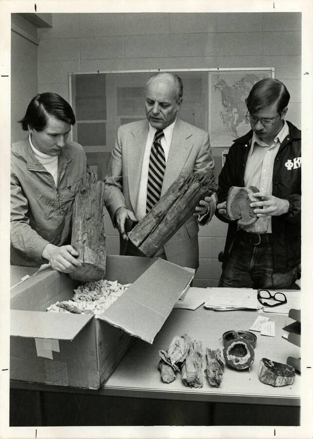 Three men looking at logs of wood on a table. One of the men is wearing a Phi Kappa jacket and another one is wearing a suit and tie.