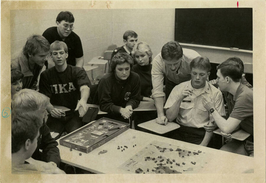 Students play Risk the board game in what looks like a classroom. Note the man on the left wearing a Pi Kappa Alpha  fraternity sweatshirt.